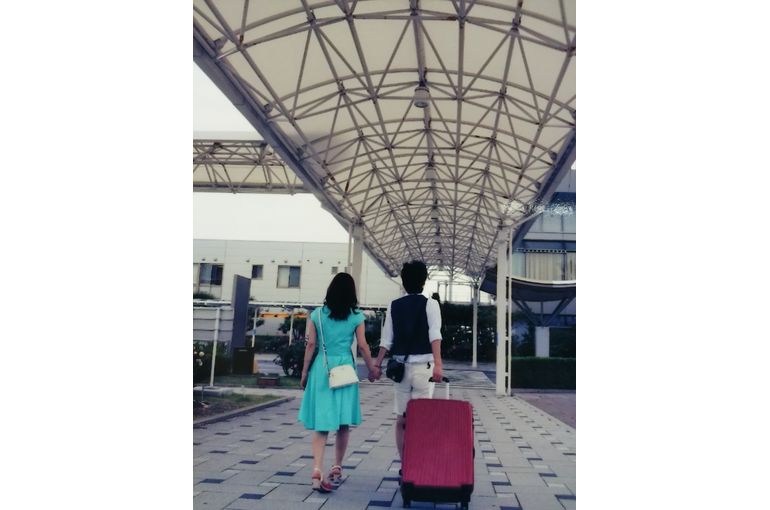 ワンピースとベストでお洒落した二人がスーツケースを引きながら空港へ向かって歩いて行く後ろ姿