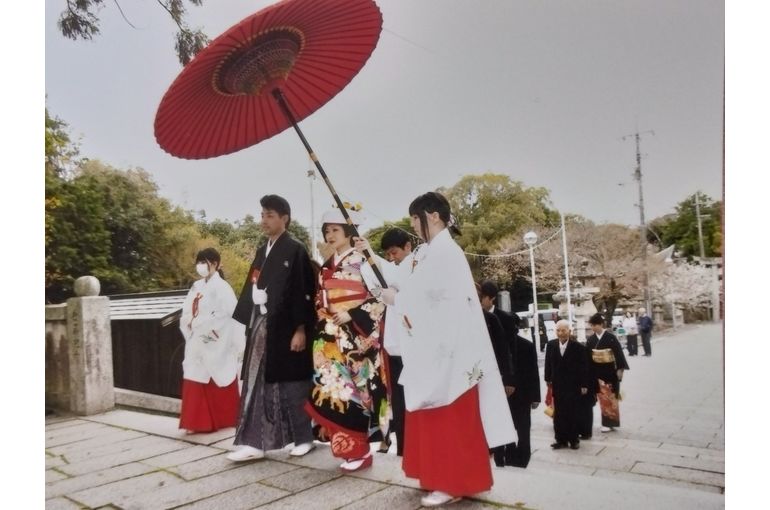 巫女が長くて大きな赤い和傘を新郎新婦に差しかけている。親族がその後ろに二列に並んで歩いている。