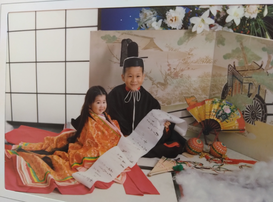 2歳の女の子と5歳の男の子が平安時代の衣装を着て、畳に座って巻紙を持ってニッコリ笑っている。
