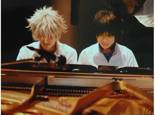 中学校男子夏の制服を着たカヲル役(銀髪)の次男が高音側、シンジ役(黒髪)の母親が低音側に並んで座り演奏する姿をピアノ越しに正面から写している