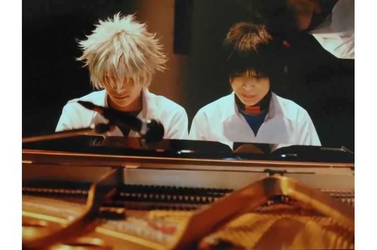 中学校男子夏の制服を着たカヲル役(銀髪)の次男が高音側、シンジ役(黒髪)の母親が低音側に並んで座り演奏する姿をピアノ越しに正面から写している